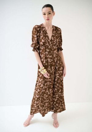 Donna in piedi con un vestito marrone a fiori con maniche a sbuffo, che indossa braccialetti dorati e tacchi rosa chiaro con cinturino, su uno sfondo bianco semplice.