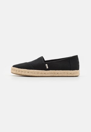 Black slip-on canvas shoe with beige braided rope sole and small side fabric tag on a white background, side view.