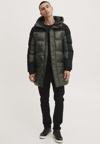 Grønn og svart dunjakke med hette, med quiltet design, glanset tekstur og frontglidelås, kombinert med svarte bukser og sko.