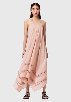 Kvinde iført en lyserød, ærmeløs, flydende kjole med blondekant og sorte, flade sandaletter med stropper, stående mod en lysegrå baggrund.