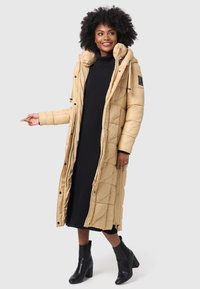 Long, puffer coat in light beige with a quilted design, featuring a high collar and zippered front. Worn over a ribbed black dress and ankle boots.