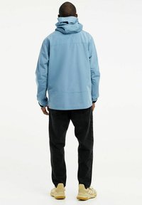 Šviesiai mėlynas vandeniui atsparus džemperis su kapišonu, ilgomis rankovėmis ir juodais akcentais, dėvimas su juodais džinsais ir geltonais sportbačiais.