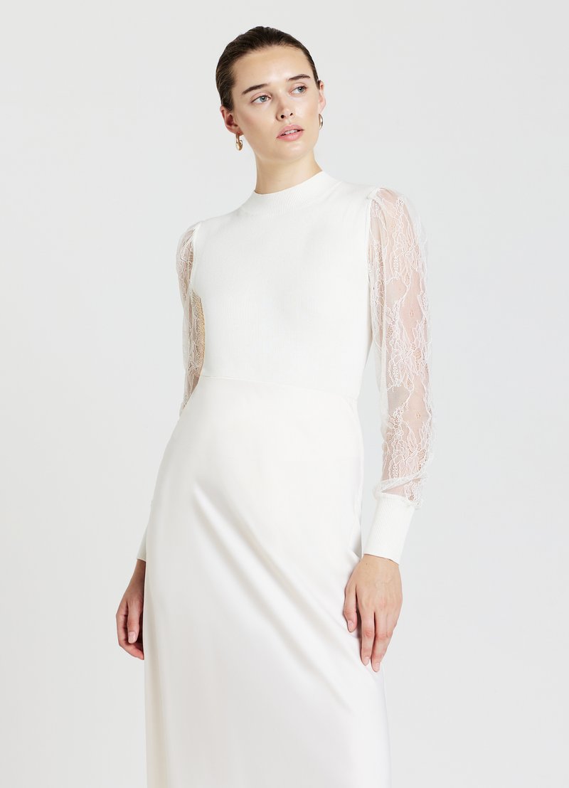 Langärmliges Kleid in Weiß, mit einer taillierten Oberseite aus strukturiertem Ripp und transparenten Spitzeärmeln, die in einen glatten, gefältelten Rock übergehen.