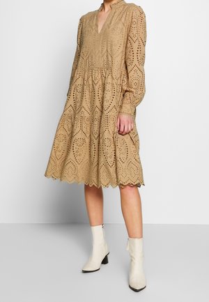 Beige oogjesjurk met lange mouwen, V-hals, gelaagde rok, ingewikkeld uitgesneden patroon en een geschulpte zoom; gecombineerd met witte enkellaarzen.