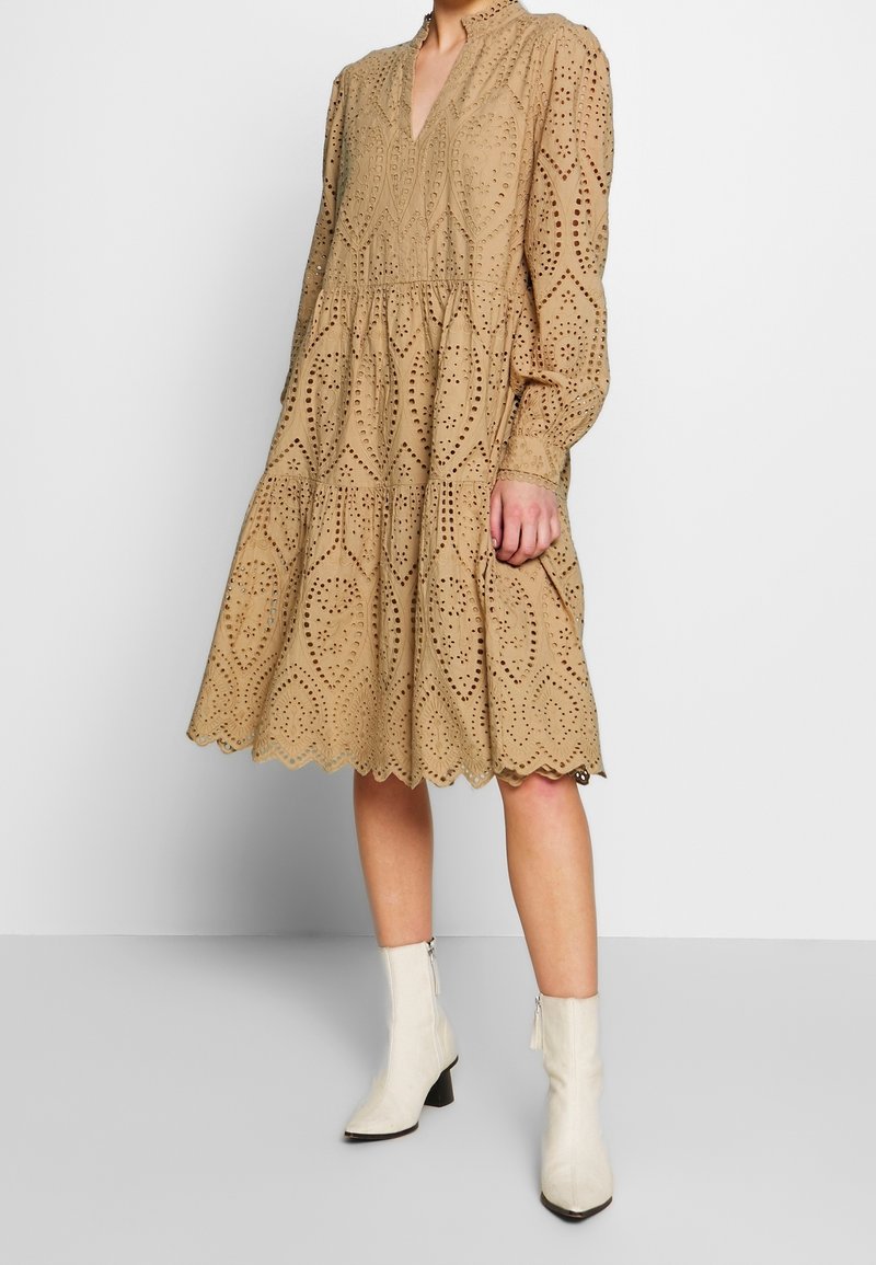 Robe beige en œillet avec manches longues, col en V, jupe à volants, motif découpé complexe et ourlet festonné ; associée à des bottines blanches.