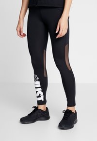 Czarne legginsy sportowe z siateczkowymi panelami, z dużym białym napisem "JUST" na lewym udzie. Połączone z czarnymi butami sportowymi.