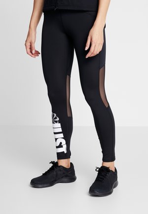 Leggings atléticos negros con paneles de malla, con una gran impresión blanca de "JUST" en la pierna izquierda. Combinados con zapatillas deportivas negras.