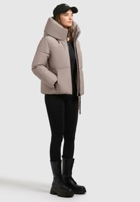 Puffjakke i lys beige med høj krave, lynlåslukning og to lommer. Sammen med sorte leggings og knæhøje snørebod.