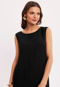 Czarna sukienka bez rękawów z okrągłym dekoltem. Gładki materiał i luźny krój. Dodatki w postaci dużych złotych kolczyków w kształcie kół.