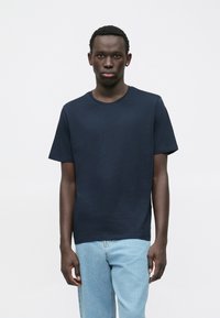 Giovane uomo con capelli corti che indossa una maglietta blu navy e jeans azzurri chiari, in piedi contro uno sfondo bianco uniforme.