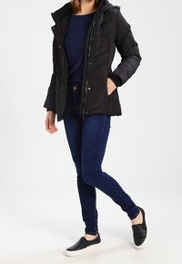 Giacca imbottita nera con cappuccio, abbinata a un top blu navy e jeans skinny blu scuro. Le scarpe slip-on nere completano l'outfit.