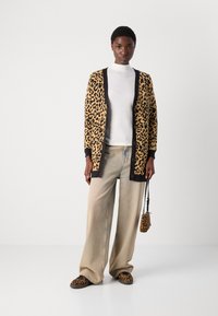 Kardigan s leopardovim vzorom v bež in črni barvi, z gumbnim zapiranjem in rebrastimi manšetami, v kombinaciji s svetlimi širokimi hlačami in usklajenimi čevlji.