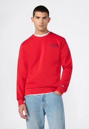 Junger Mann trägt ein rotes Champion-Sweatshirt mit Logo auf Brust und Ärmel, kombiniert mit hellblauen Jeans, stehend vor einem schlichten Hintergrund.