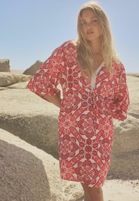 Kaftan à motifs rouge et blanc avec une coupe décontractée, des manches larges et des bords en dentelle. Un design floral texturé couvre le tissu.