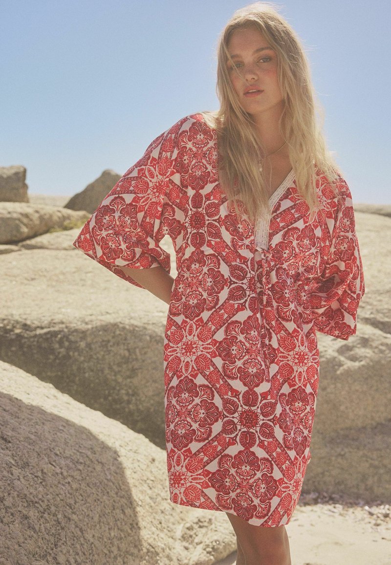 Kaftan à motifs rouge et blanc avec une coupe décontractée, des manches larges et des bords en dentelle. Un design floral texturé couvre le tissu.
