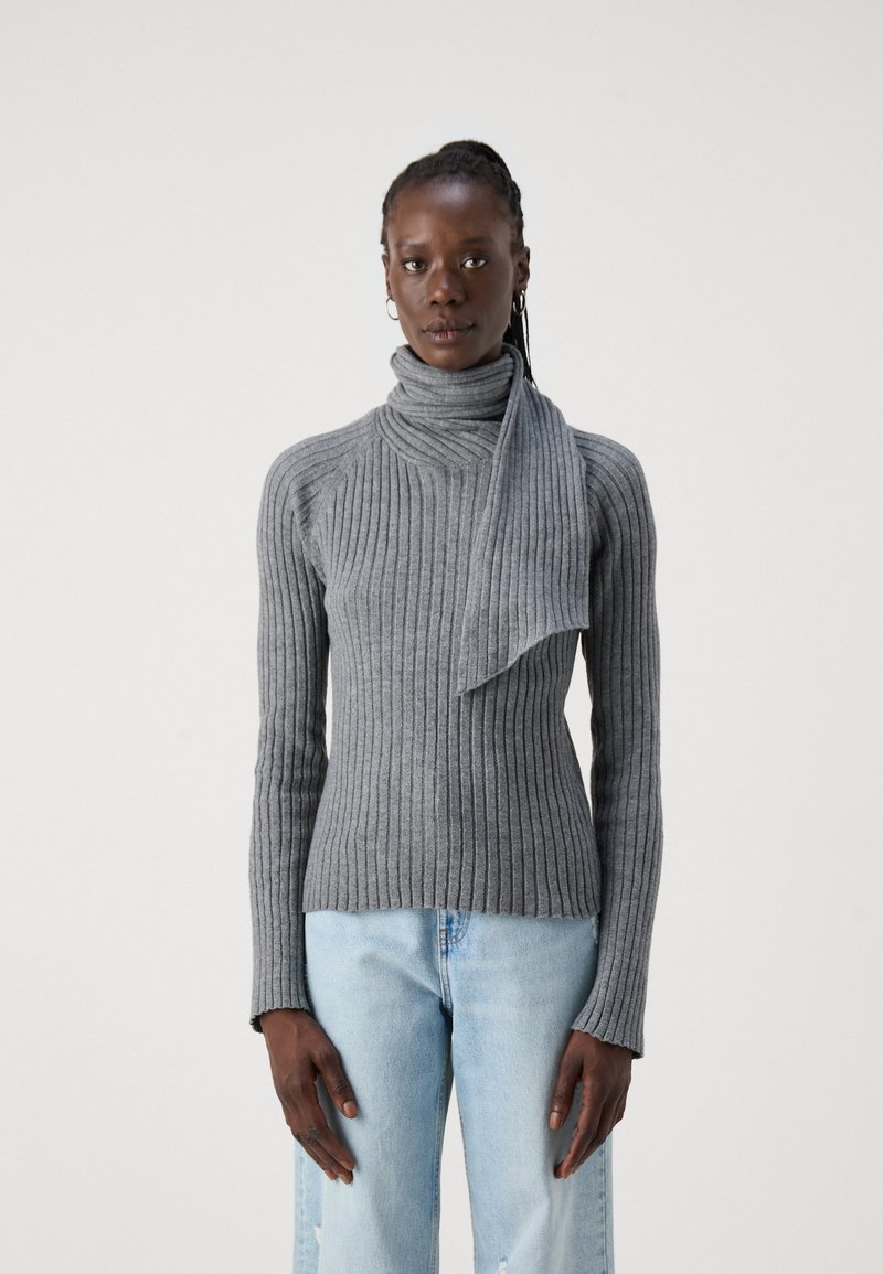 Sivi rebrasti pulover s turtleneck ovratnikom in dolgimi rokavi. Kombiniran z svetlo modrimi hlačami, ki imajo sproščen kroj.