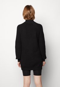 Pull en maille noire avec un col montant, des manches longues et une texture côtelée, affichant une coupe décontractée et des fentes sur les côtés au niveau de l'ourlet.