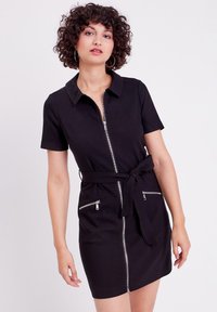 Vestido negro con cremallera, mangas cortas, cuello y cinturón en la cintura. Cuenta con dos bolsillos frontales con cremallera y una tela texturizada.