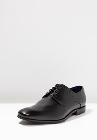 Chaussure de ville en cuir noir présentant un design élégant, une pointe arrondie, une fermeture à lacets et une texture subtile avec un talon empilé bas.