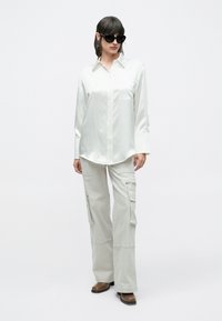 Femeie purtând o cămașă albă din satin supradimensionată, pantaloni cargo bej, cizme maro și ochelari de soare negri, pe un fundal simplu și deschis la culoare.