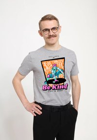 Šedé bavlněné tričko s barevným grafickým designem, na kterém je skateboardista a fráze "Buď hodný" v růžové barvě, umístěná na živém oranžovém pozadí.