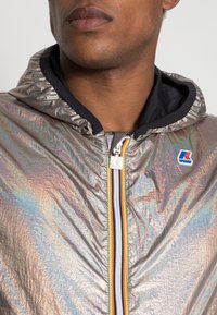 Iridescente windbreaker met capuchon, ritssluiting aan de voorkant en contrasterende gestreepte ritssluiting. Heeft een subtiele logo op de borst. Gladde, reflecterende textuur.