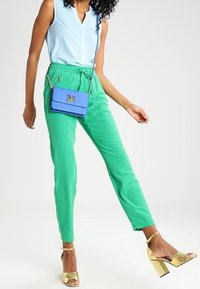 Grønne bukser med elastisk liv, parret med en lyseblå ærmeløs top og gyldne hæle sandaler. En lille blå crossover taske tilføjer kontrast.