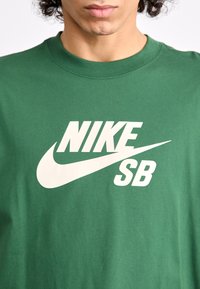 Green cotton t-shirt featuring a large white Nike SB logo and swoosh design, round neckline, and short sleeves with a smooth texture.