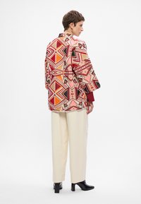 Mintázott dzseki élénk színekben, geometrikus mintákkal, krémszínű nadrággal párosítva. Lazán illeszkedő fazon, sálgallérral.