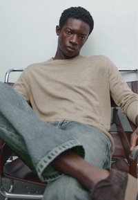Beige knit sweater, relaxed fit; light blue jeans with frayed cuffs; brown slip-on shoes; seated on a chair with leather accents.