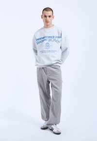Grijze sweatshirt met blauwe grafische tekst en een wereldbolontwerp, gecombineerd met lichtgrijze katoenlook broek en sneakers in neutrale tinten.