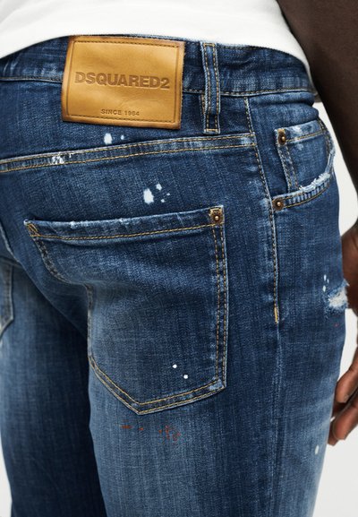 Gros plan sur un jean bleu délavé avec des éclaboussures de peinture et un patch en cuir tan DSQUARED2 sur la ceinture.
