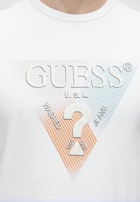 Λευκό t-shirt με μεγάλο λογότυπο Guess σε τρίγωνο, που περιλαμβάνει το κείμενο "GUESS U.S.A." και ένα μεγάλο ερωτηματικό στο κέντρο.