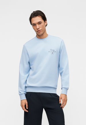 Mand iført en lyseblå Tommy Hilfiger sweatshirt med logo på brystet og mørke bukser, stående mod en hvid baggrund.