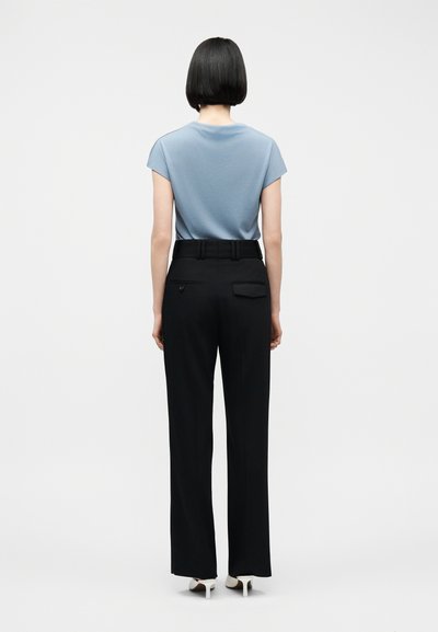 Moters trumpi juodi plaukai, vilkinti šviesiai mėlyna trumpomis rankovėmis palaidine, įkišta į aukštu juosmeniu juodas kelnes, su baltomis aukštakulnėmis, matoma iš nugaros.