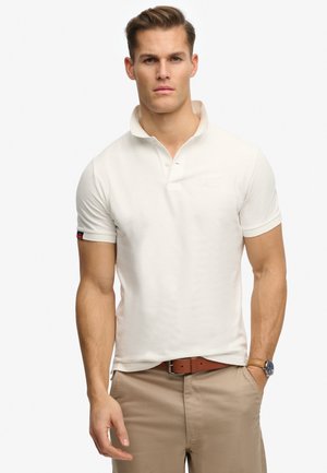 Homme portant un polo à manches courtes couleur crème, pantalon beige, ceinture marron et montre, debout avec une main dans la poche sur un fond uni.