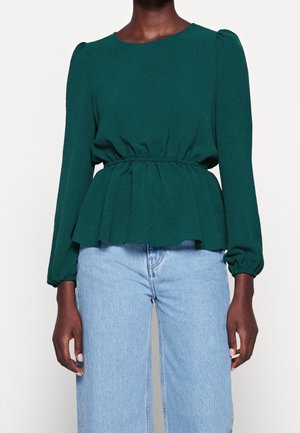 Vrouw die een donkergroene blouse met lange mouwen en een gerimpelde taille draagt, gecombineerd met lichtblauwe, hooggetailleerde jeans, tegen een effen achtergrond.