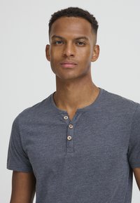 Camisa henley de manga corta en gris oscuro con tres botones de madera en el escote, hecha de una tela suave y texturizada. Diseño simple y limpio.
