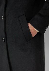 Schwarzer Wollmantel mit Knopfverschluss, glatter Textur und seitlichen Taschen, der ein tailliertes Design zeigt.