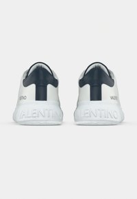 Λευκά αθλητικά παπούτσια με μαύρες ετικέτες στη φτέρνα, λογότυπο Valentino στις εξωτερικές πλευρές, και την επιγραφή «VALENTINO» χαραγμένη στις παχιές λευκές σόλες, εμφανιζόμενα από πίσω.