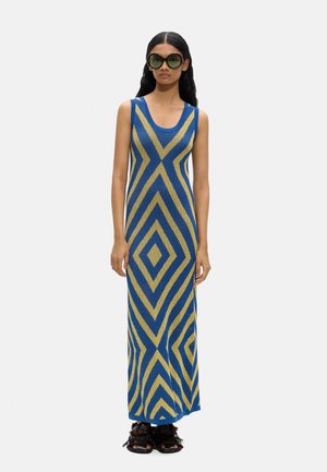 Kobieta w niebiesko-żółtej, geometrycznie wzorzystej sukience maxi bez rękawów, duże okrągłe okulary przeciwsłoneczne oraz czarne sandały z frędzlami.