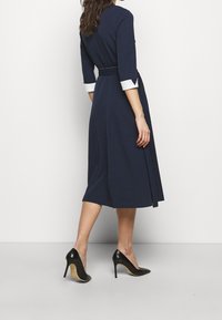 Navyblaues Kleid mit dreiviertel Ärmeln und weißen Manschetten, tailliertem Bund, schwingendem Rock und kombiniert mit schwarzen High Heels.