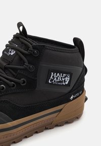 Svart og grå mid-top sneaker laget av semsket skinn og mesh-materialer, med gummisåle, snøring og en "Half Cab" logo-patch.