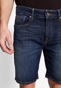 Shorts de mezclilla en azul oscuro con una cintura de altura media, cinco bolsillos y costuras naranjas en contraste. Dobladillo enrollado en la parte inferior.