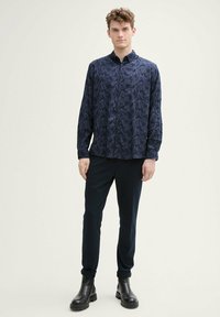 Donkerblauwe overhemd met lange mouwen, bladpatroon, knoopsluiting, zachte stof; gecombineerd met donkere broek en zwarte laarzen.