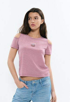 Mujer con blusa de rayas rosas y blancas con hombros descubiertos y un pequeño gráfico, combinada con jeans de mezclilla azul claro, posando con una mano en el bolsillo.