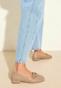Beige flat shoes with a rounded toe, shiny finish, and a decorative gold chain accent. Paired with light blue cuffed jeans.