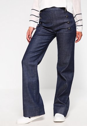 Kobieta ubrana w ciemnoniebieskie, szerokie jeansy z wysokim stanem i ozdobnymi guzikami po bokach, białe trampki oraz białą bluzkę z czarnymi paskami na rękawach.