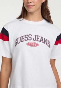 Biela bavlnená tričko s červenými a námorníckymi pruhmi na rukávoch. Predný grafický nápis "GUESS JEANS 1981" v tučnom, textúrovanom písmene.
