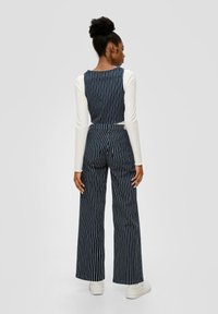 Gestreifter Denim-Overall mit weitem Bein und taillierten Oberteil, Rückansicht, in dunkelblau mit weißen Streifen. Kombiniert mit einem weißem Langarmoberteil.